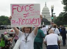 A NCIL Member holds a sign that reads "Force Does Not Equal Recovery"