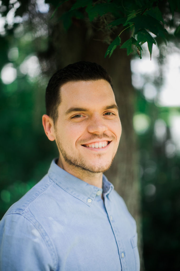 A young white man wearing a blue button-up shirt smiles at the camera