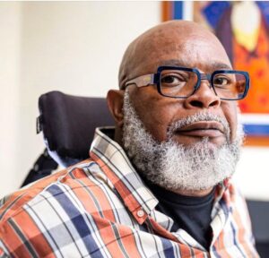 Theo Braddy Headshot - A black man with a bald head, a salt and pepper full beard wearing eyeglasses, sitting in a wheelchair wearing an orange, blue, and white plaid dress shirt.